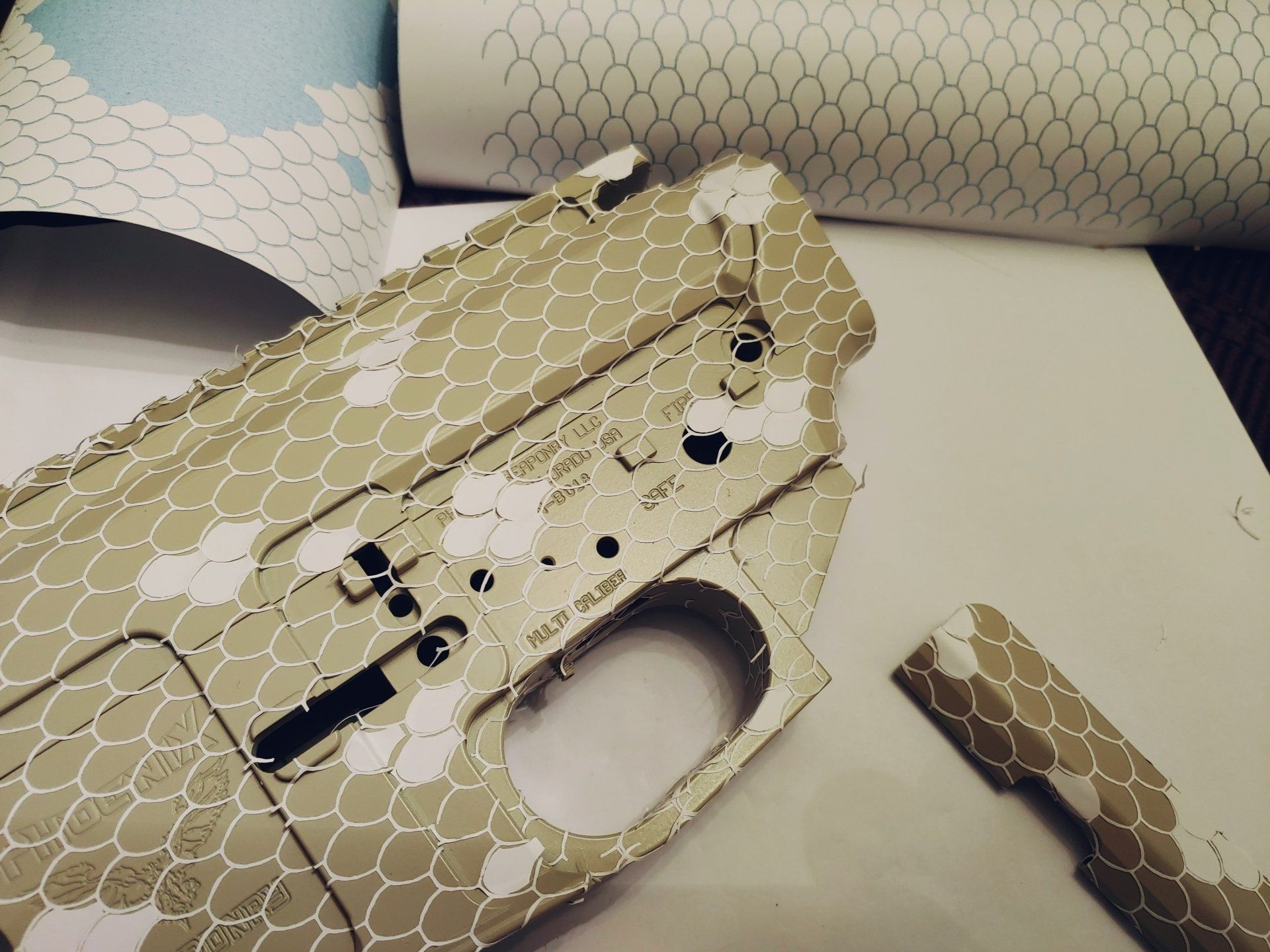 Tan and white camouflage pattern on firearm components with rolls of patterned material in the background.