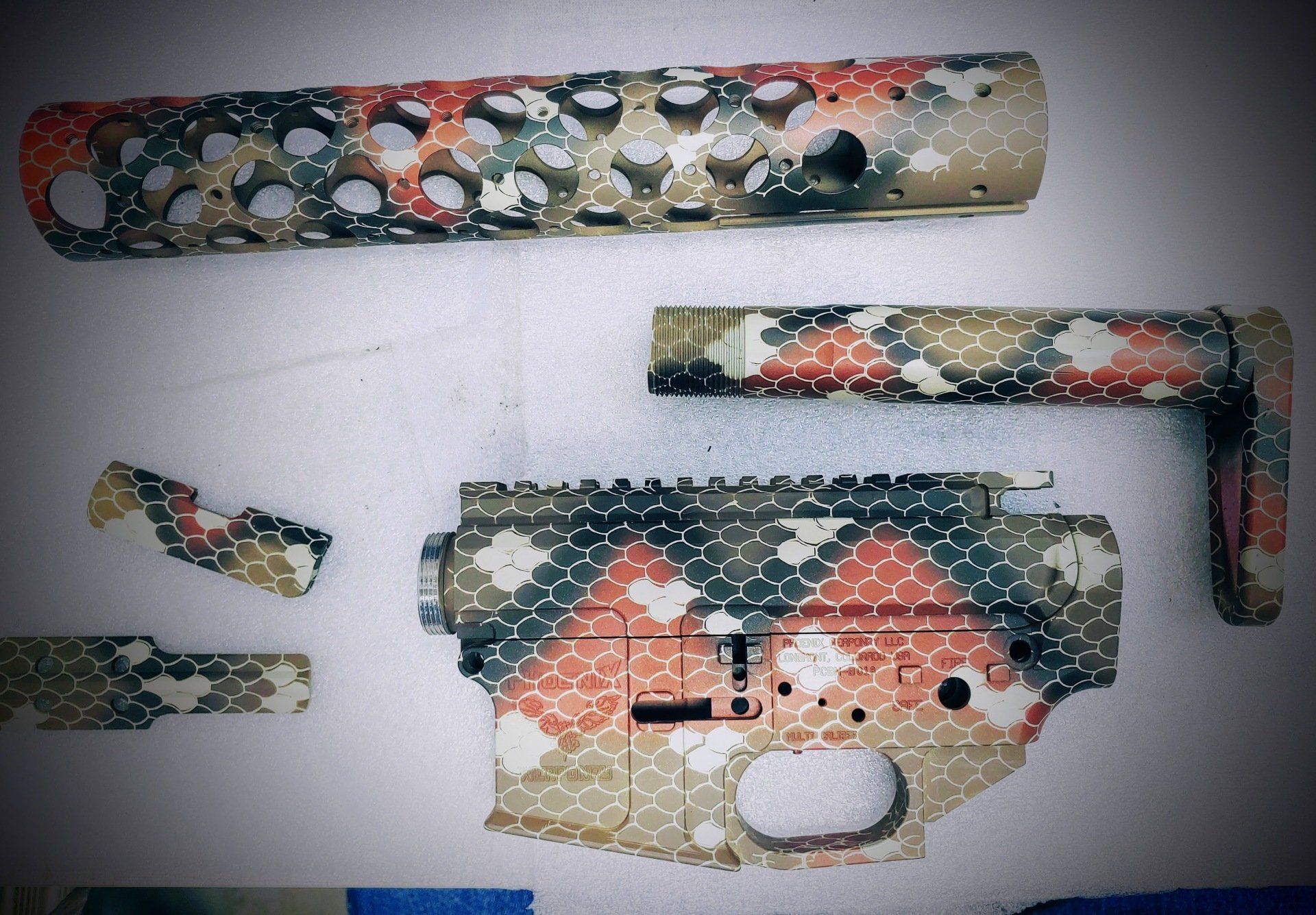 Camouflage-painted firearm components: handguard, stock, lower receiver, various parts. Tan, red, and green colors.