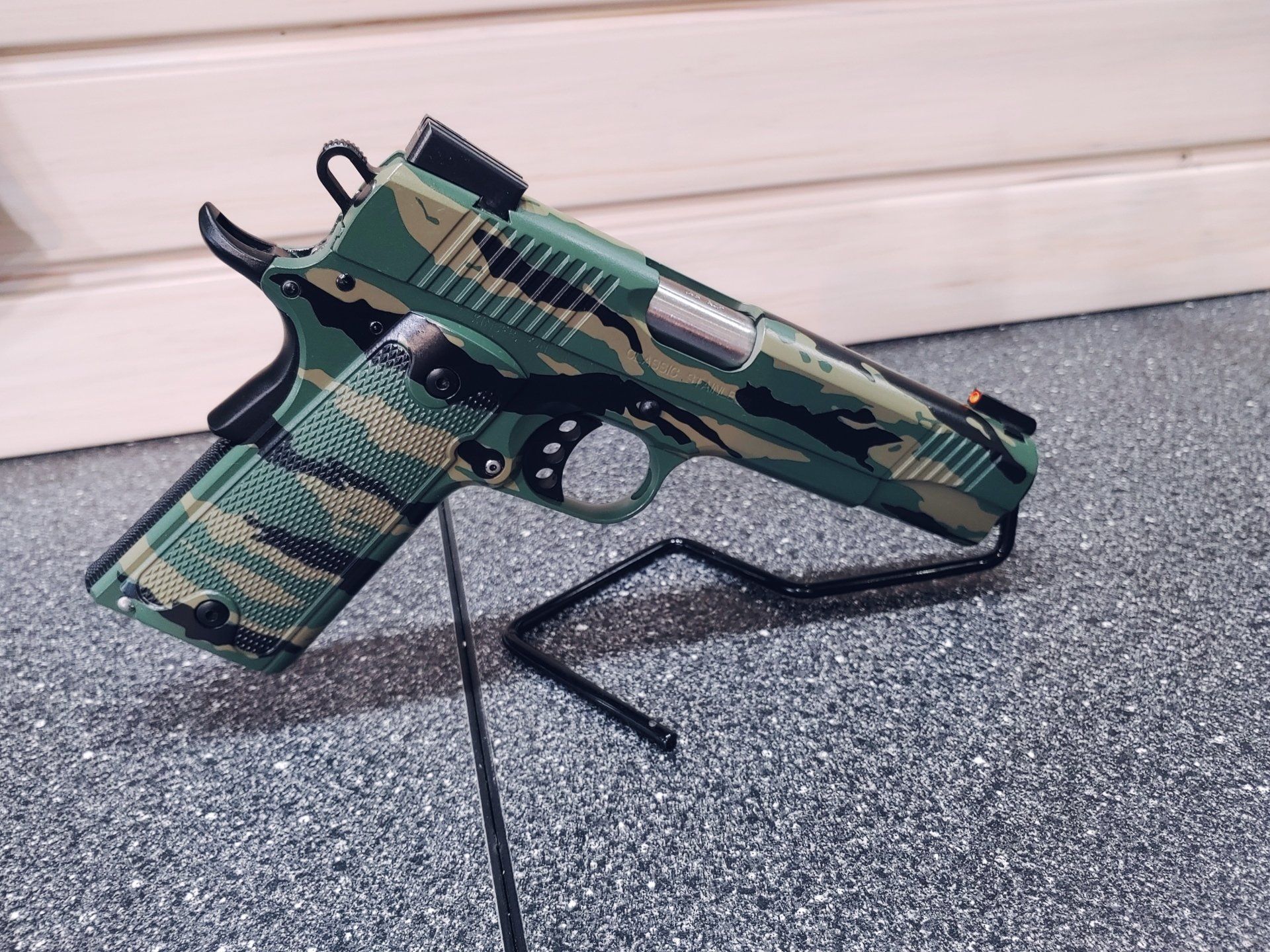 Green camouflage handgun on a black display stand against a speckled surface.