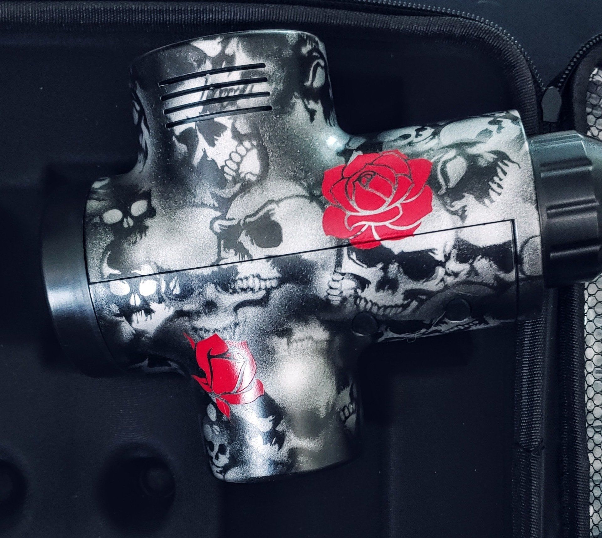 Skull-patterned massage gun with red rose accents, resting in a black case.