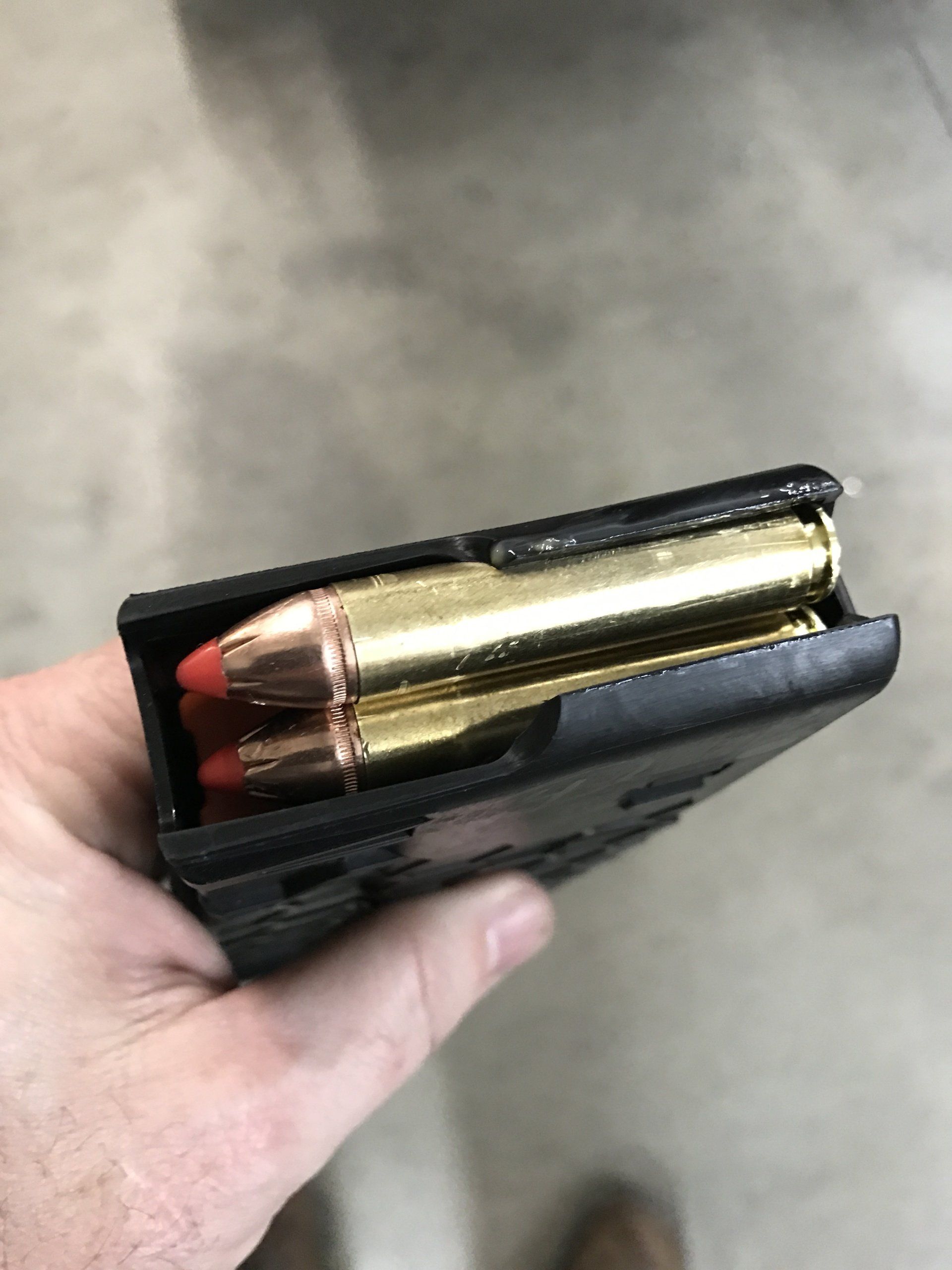 Hand holding a black firearm magazine loaded with two rifle cartridges, visible copper jackets and red tips.
