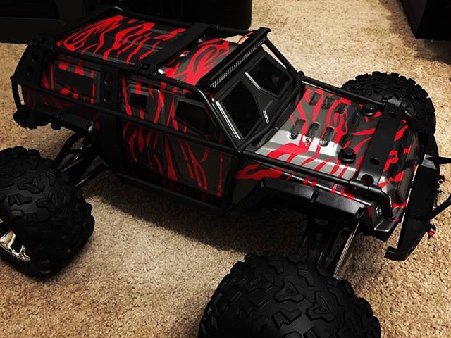 Black and red camouflage RC monster truck on a brown rug, with large tires.