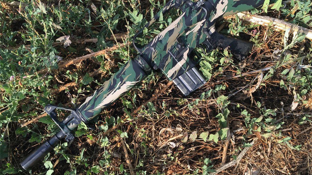 Camouflage rifle lying on a bed of green and brown foliage.