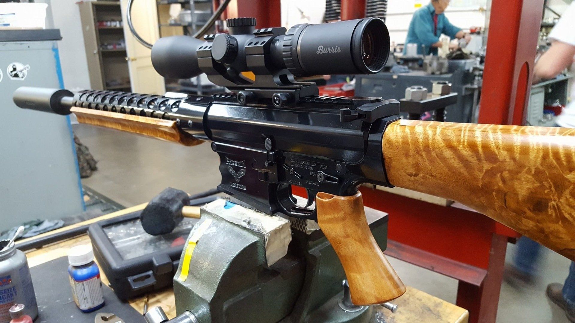 Black rifle with wooden handguard and stock, scope, and suppressor in a workshop.