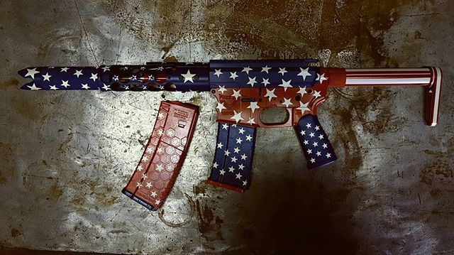 AR-15-style rifle and magazine with red, white, and blue American flag design, resting on a textured metal surface.