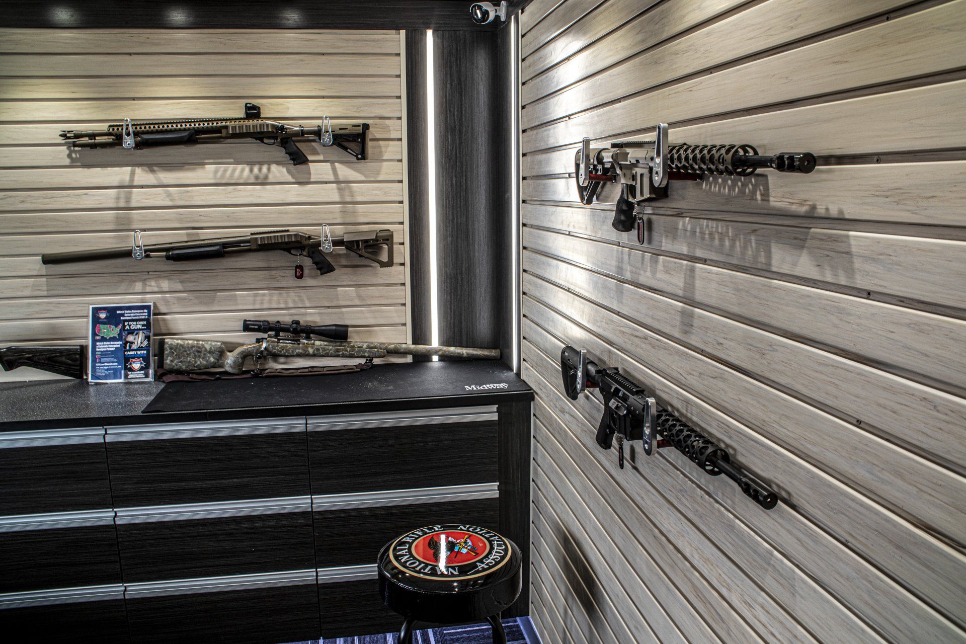 Guns displayed on a white wall in a showroom, black cabinet below, with a stool.