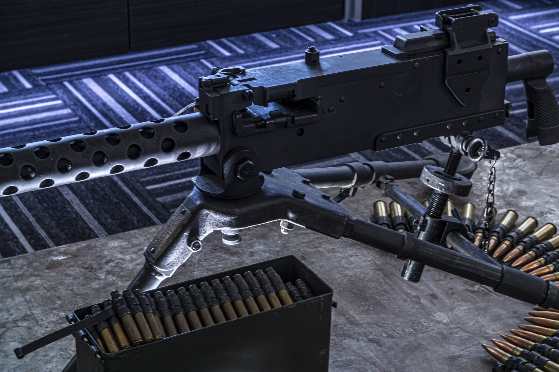 Heavy machine gun on a tripod, with ammunition belt and box, on a gray surface.