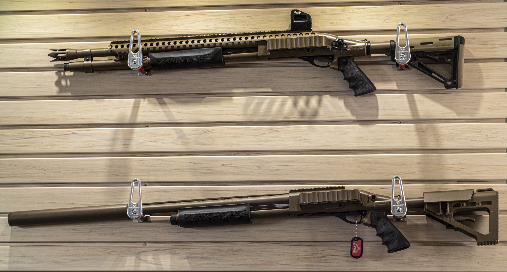 Two tan shotguns mounted on a wooden wall, one with a red dot sight and the other with a silencer.