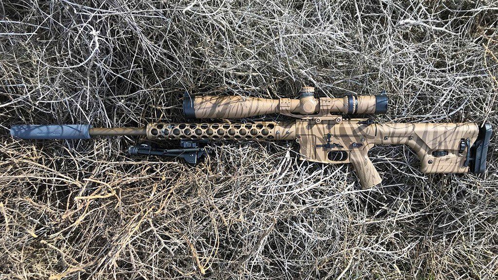 Camouflaged sniper rifle with scope and silencer on dry, grassy ground.