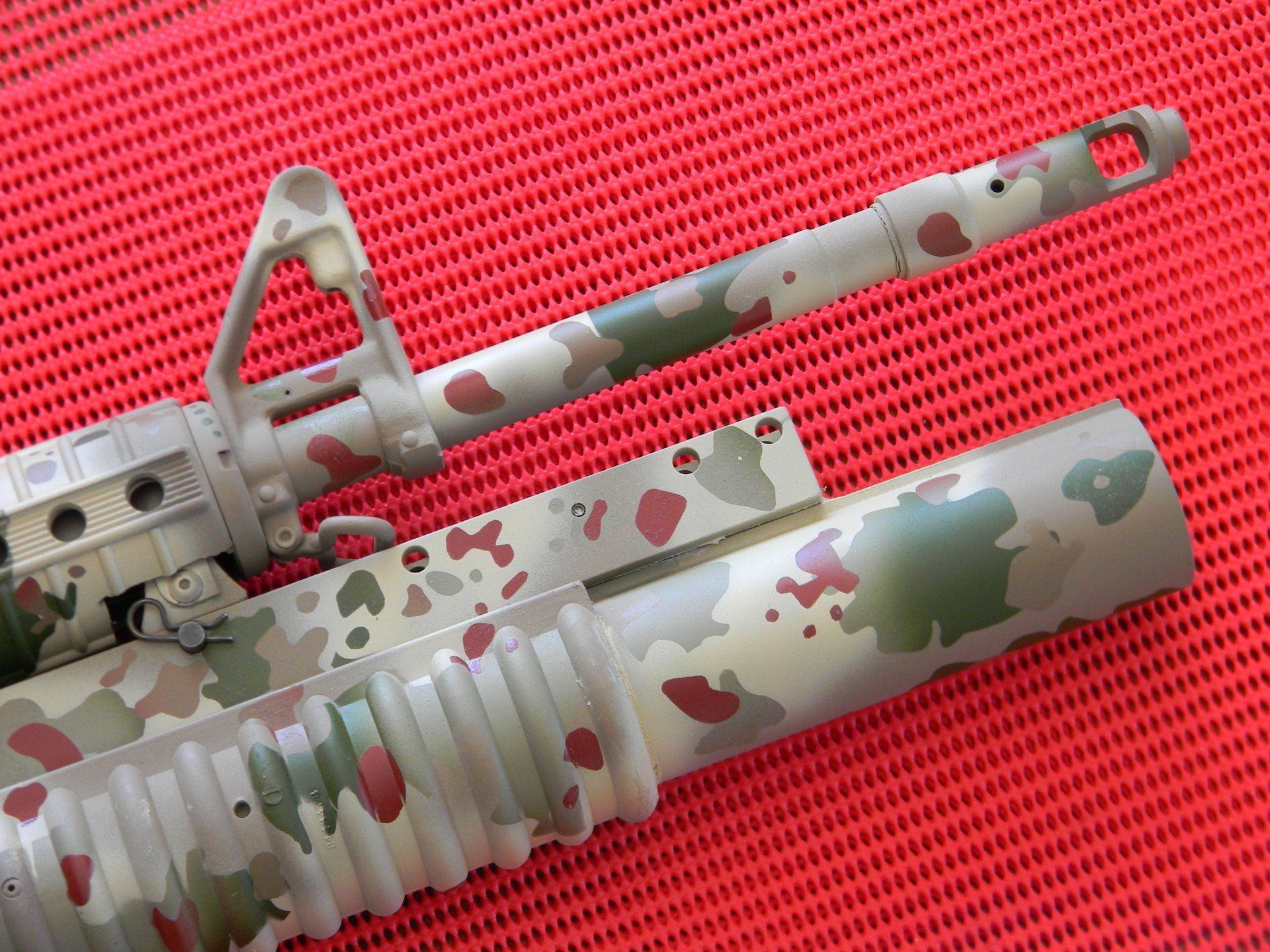 Camouflaged rifle barrel and handguard, with green, brown and white paint on red background.