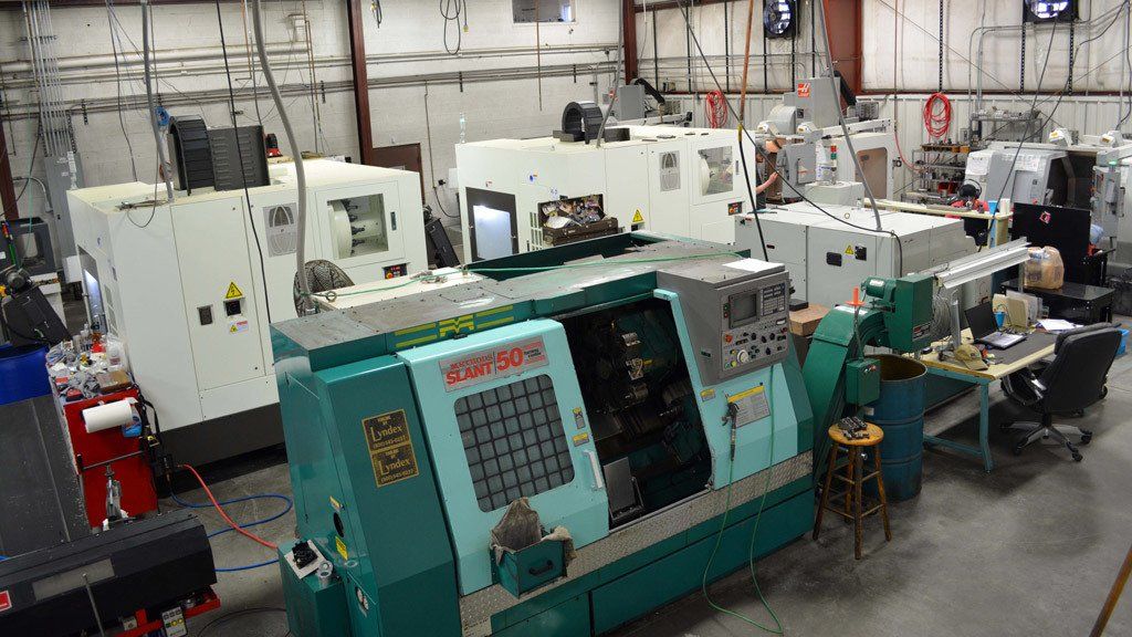 A workshop with several white and teal CNC machines, with a workstation in the background.
