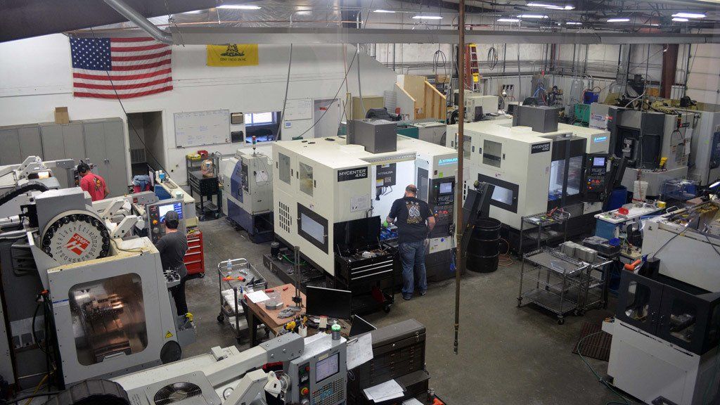 Machine shop interior with multiple computer numerical control (CNC) machines and workers. American flag visible.