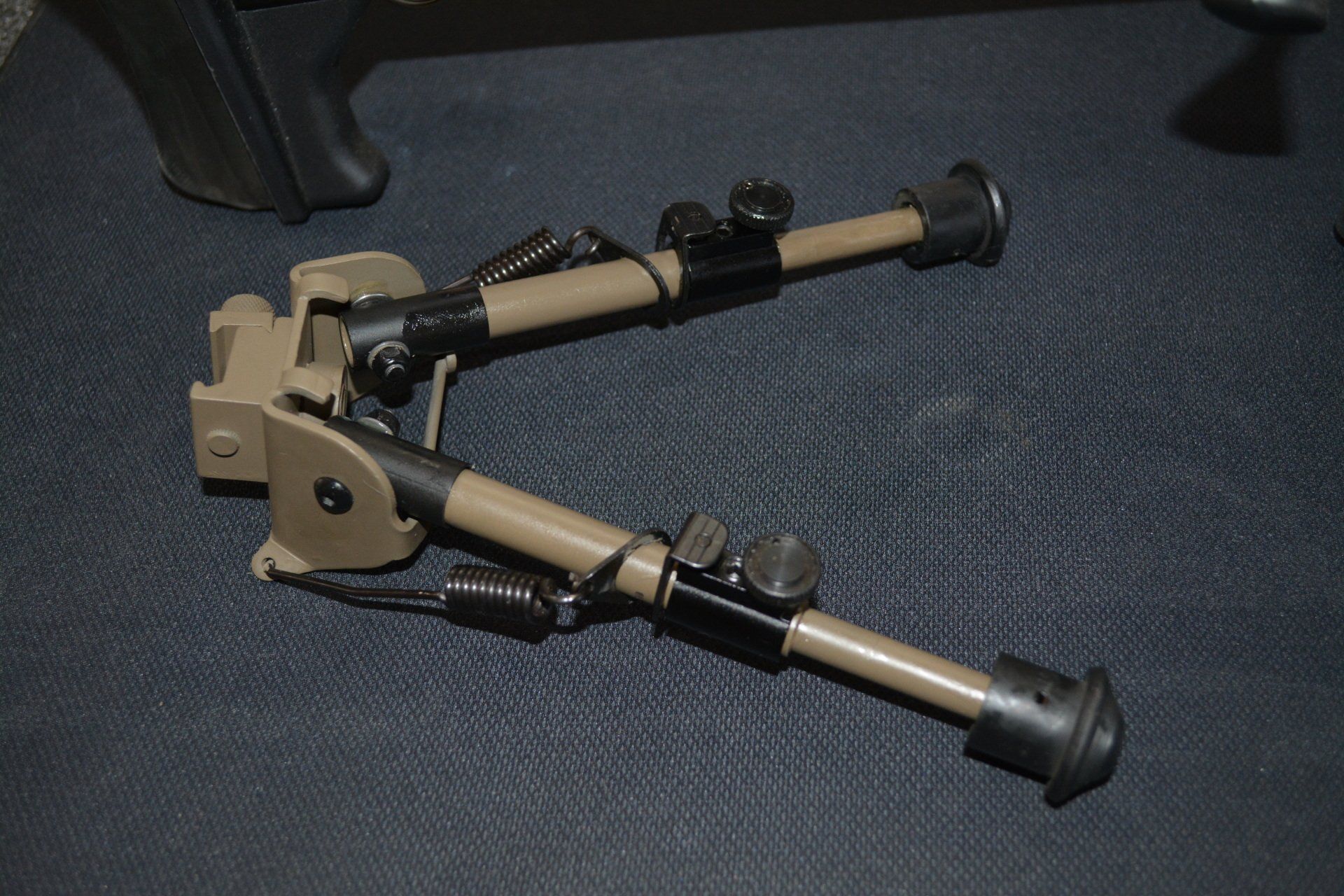 Tan and black bipod, legs extended, on a dark surface.