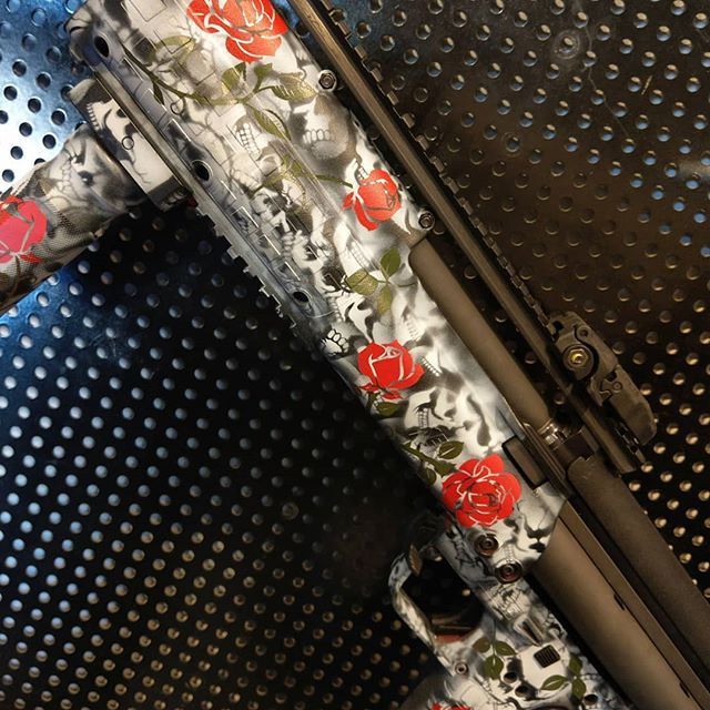 Shotgun with white, black, and gray skull-and-rose pattern against a metal backdrop. Red roses are prominent.