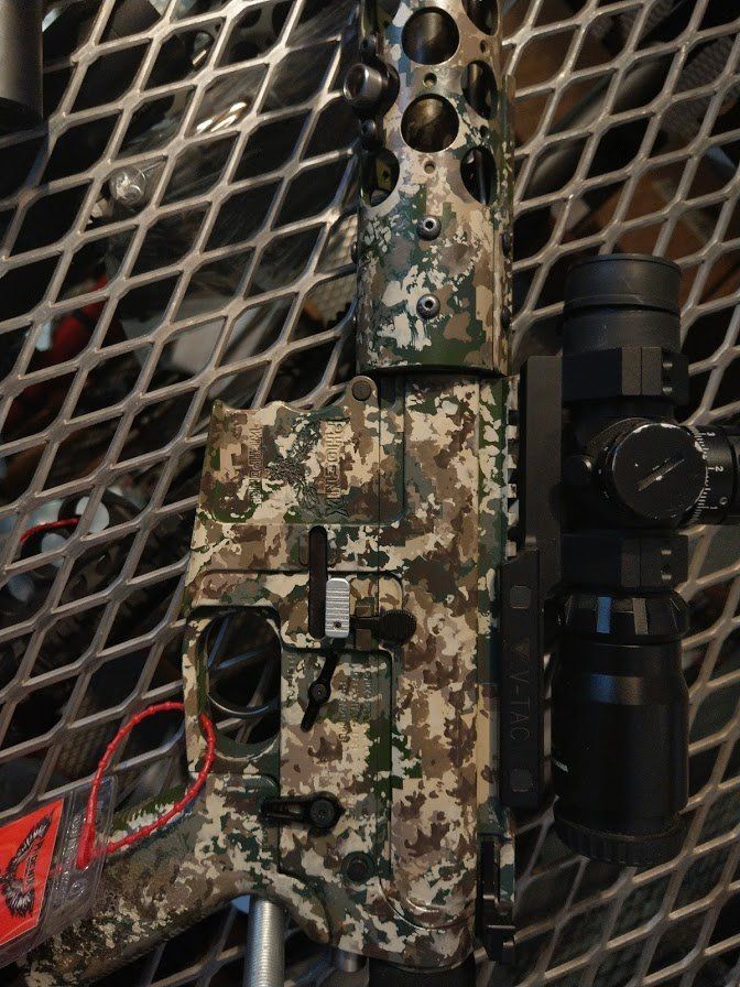 Camouflaged rifle with scope, set against a metal grid.