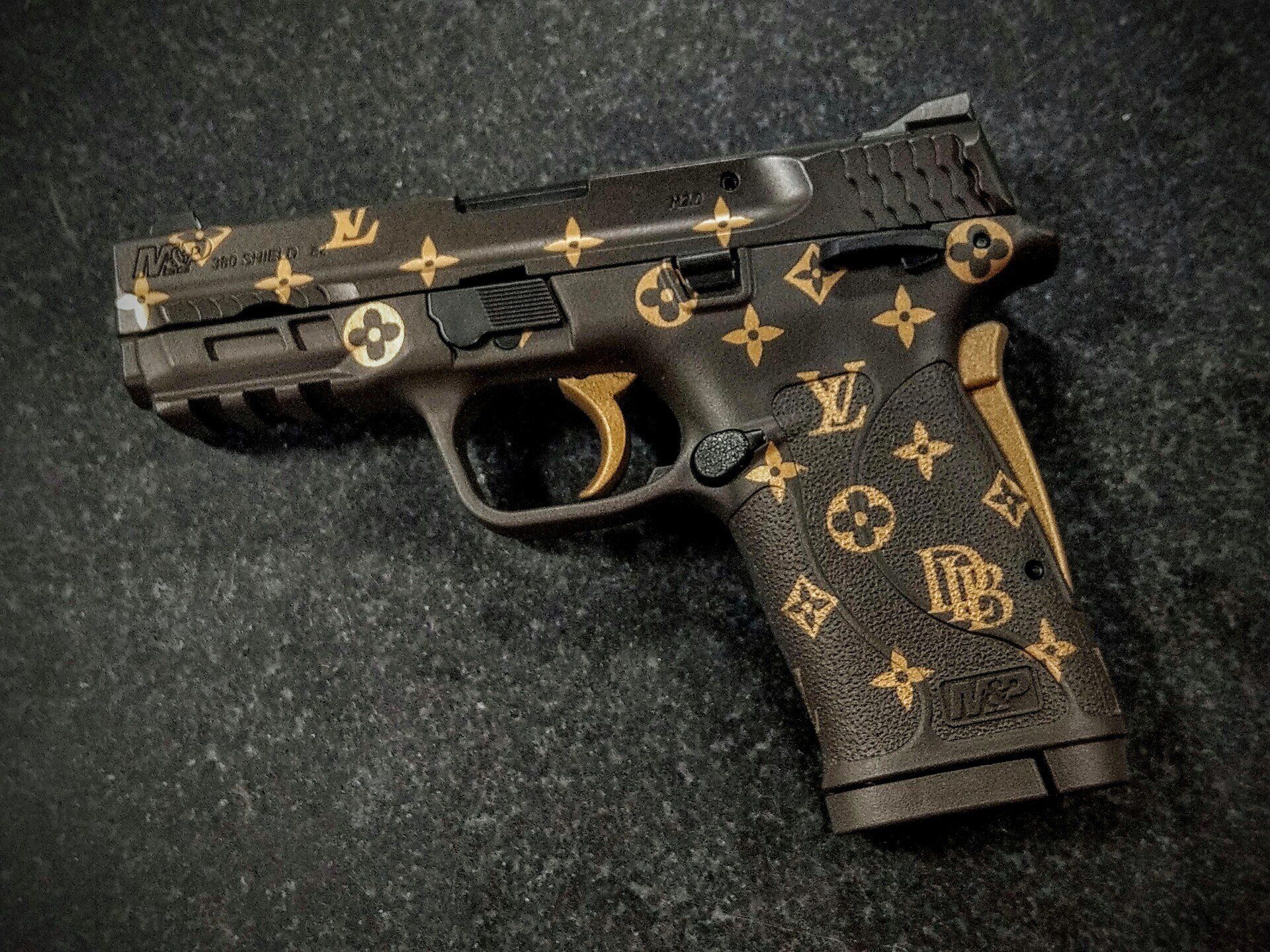 Handgun with a brown and gold Louis Vuitton pattern on a dark background.