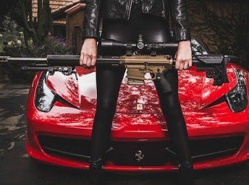 Person in black leather jacket and pants holding a rifle in front of a red Ferrari.