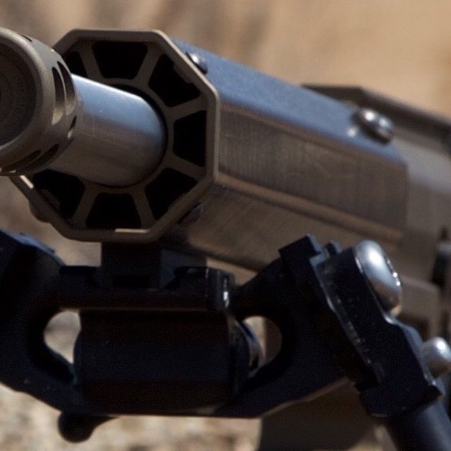 Close-up of a dark metal rifle barrel with a multi-faceted design and a bipod attached.