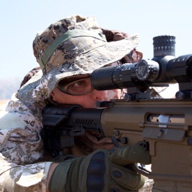 Person aiming a rifle through a scope, wearing camouflage, in an outdoor setting.