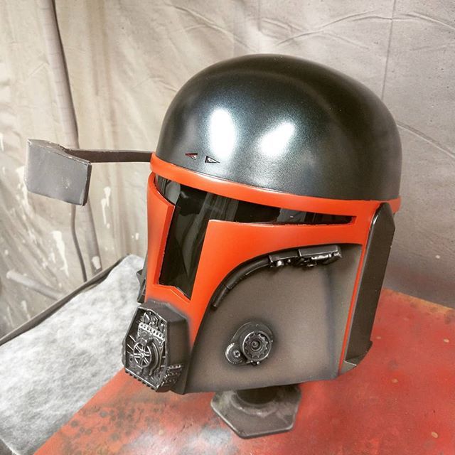 A Mandalorian helmet in shades of orange, gray, and black, with a visor, on a red surface.