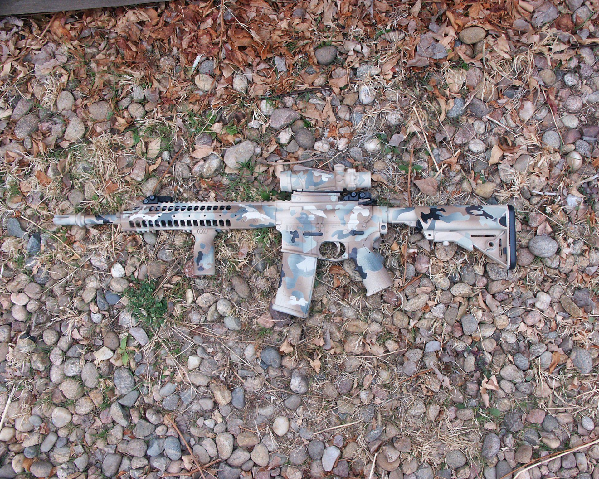Camouflage rifle lying on a bed of rocks and leaves.