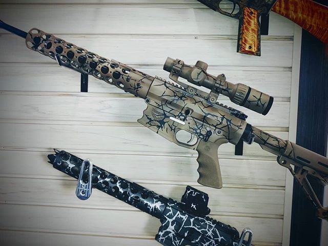 Tan and black camouflage rifle with scope mounted, displayed on a wall with other firearms.
