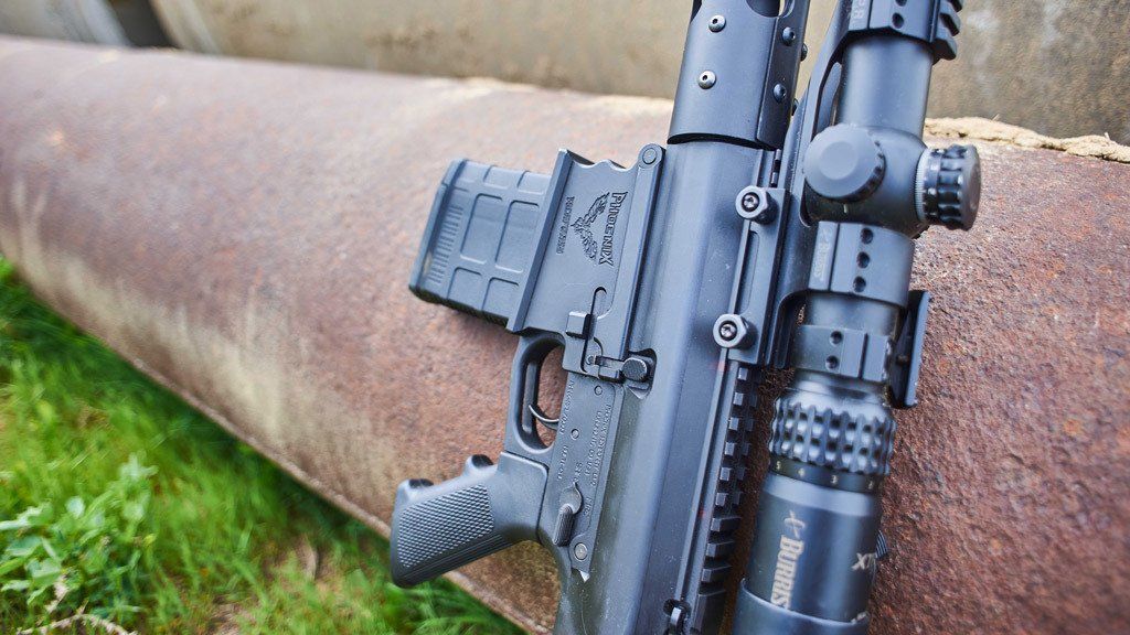 Black rifle with scope and magazine rests against a rusty metal pipe.