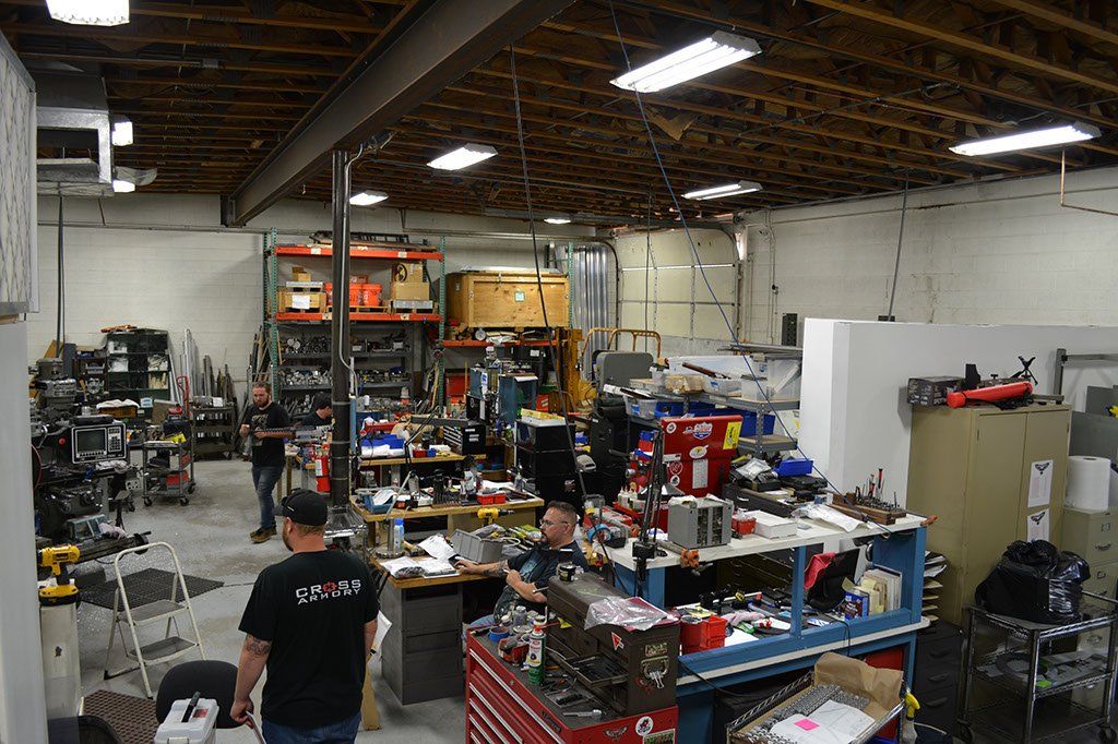 Workshop interior with several workbenches, tools, and people. Ceiling lights illuminate the space.