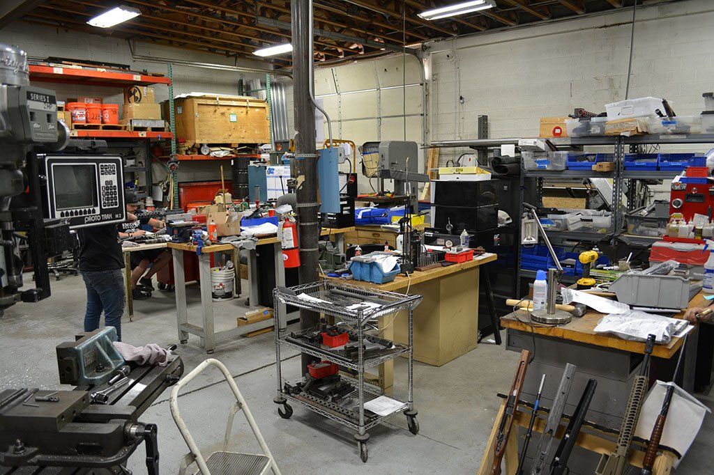 A cluttered workshop with tools, shelves, and workbenches. Several people are present, working in the space.