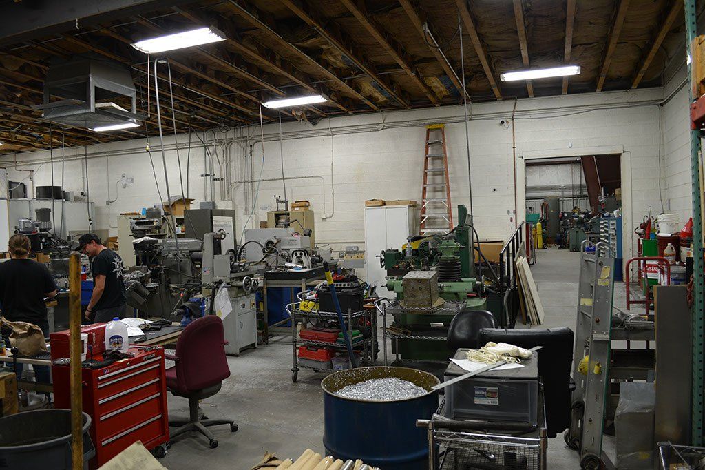 Workshop interior with machinery, tools, and people. A ladder leans against the wall, open door in the background.