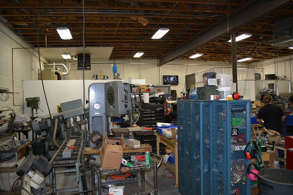 Cluttered workshop interior with machinery, tools, and a person working among shelves and containers.