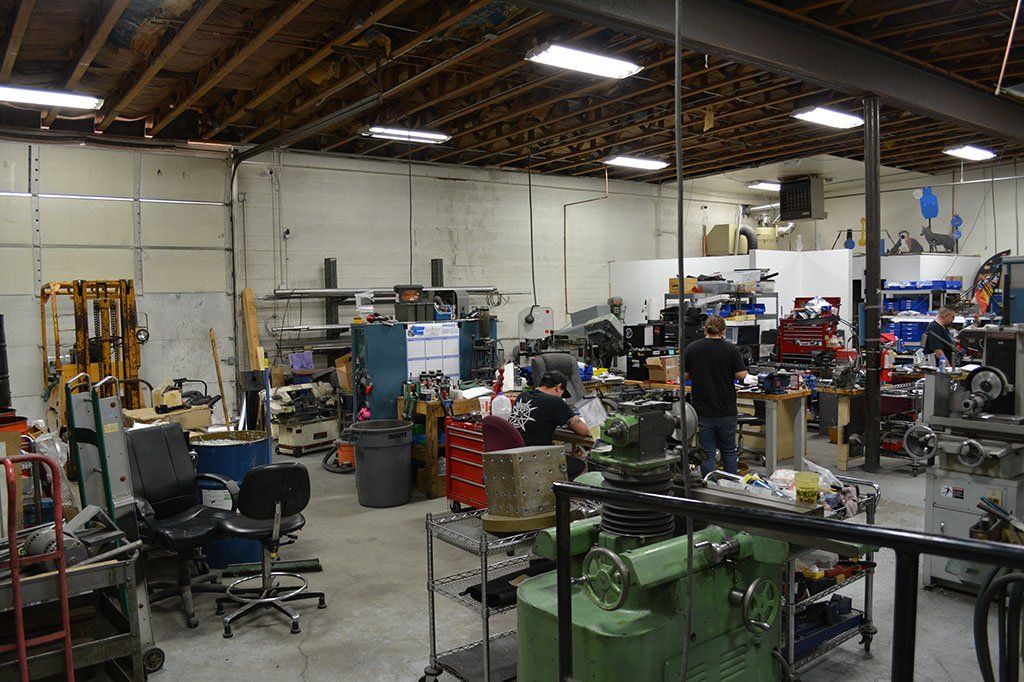Workshop interior, cluttered with machinery, tools, and people. Ceiling lights illuminate the space.