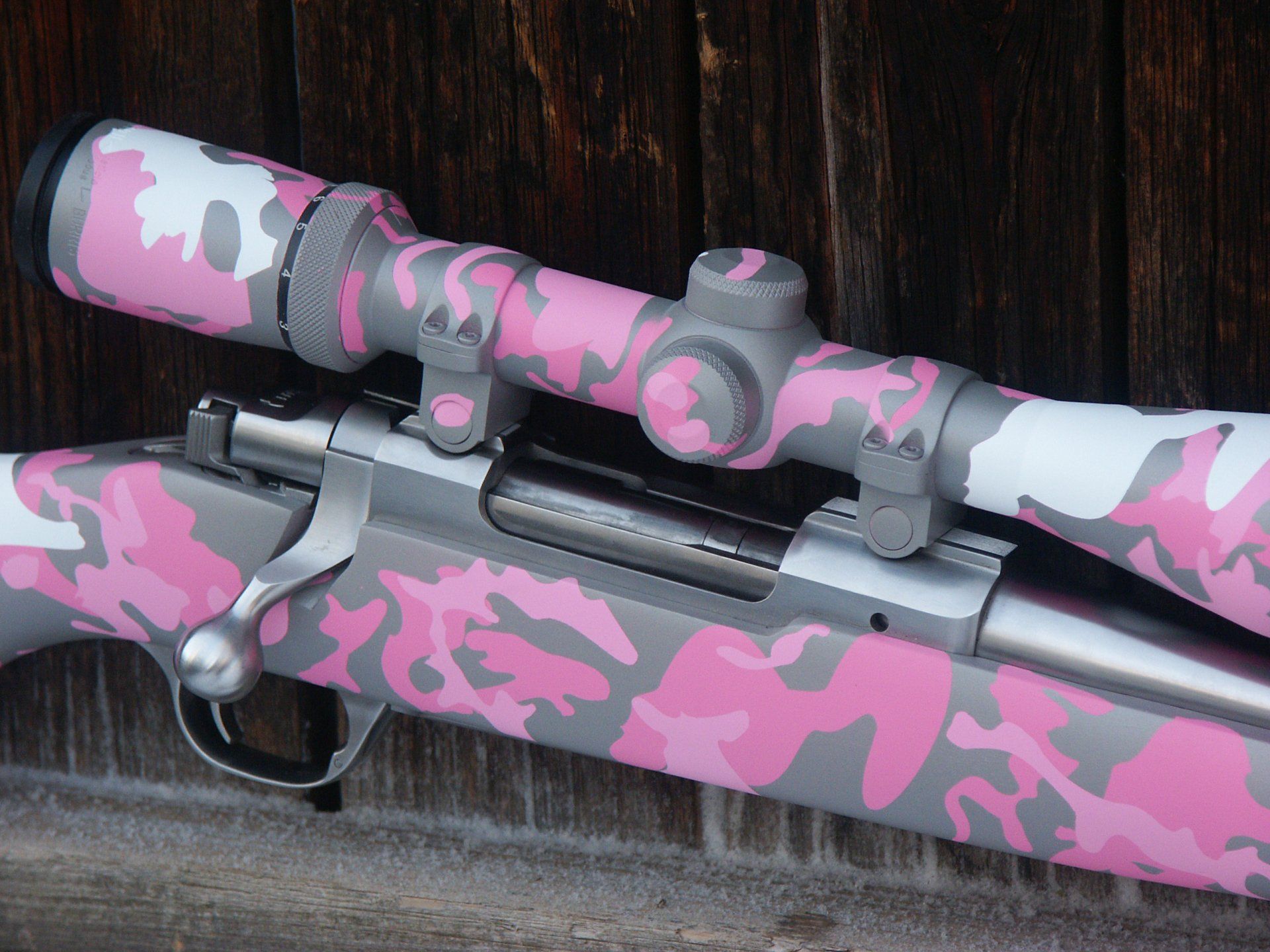 Pink and gray camouflage-painted rifle with scope, set against a wood backdrop.