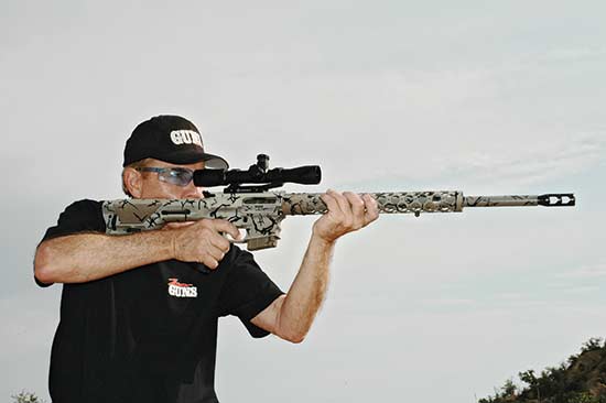 Man aiming a camouflage-patterned rifle with a scope, wearing a hat and sunglasses, against a cloudy sky.