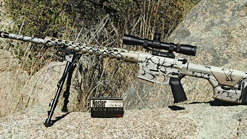 Camouflaged rifle with scope and bipod rests against rock backdrop.