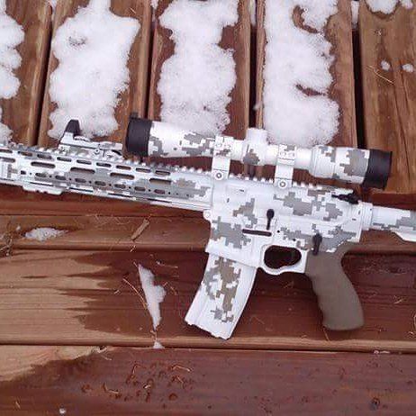 AR-15 rifle with white and gray digital camouflage pattern, scope, and magazine; resting on wood with snow.