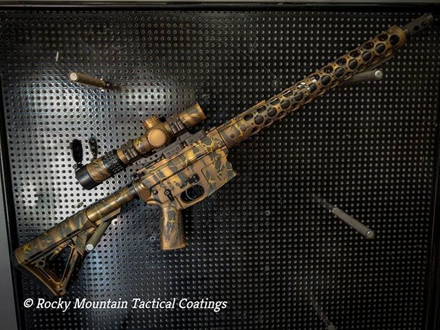 Black rifle with camouflage paint, scope, and perforated barrel, displayed on a black background.