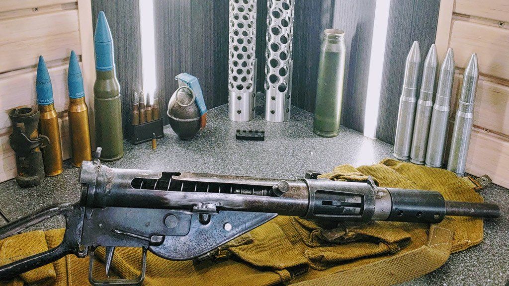 Submachine gun with ammunition and grenade on a surface with textured fabric. Various shell casings and tubes are behind.