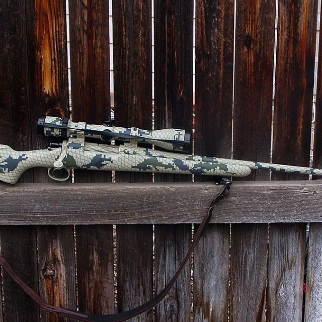 Camouflage rifle with scope, leaning against a weathered wooden fence.