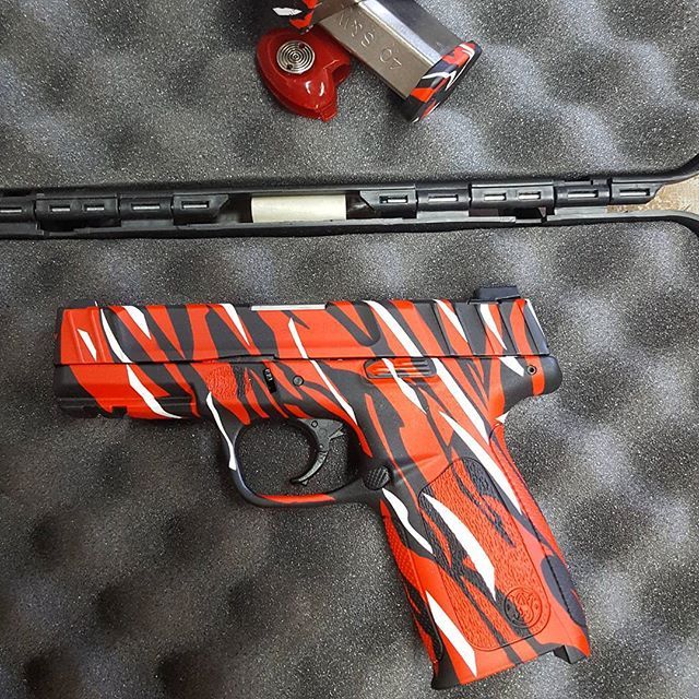 A red, black, and white camouflage handgun is in a case with a matching magazine.