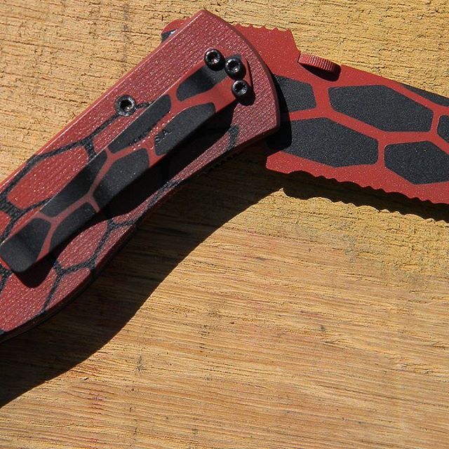Red and black folding knife on wooden surface; black honeycomb design.
