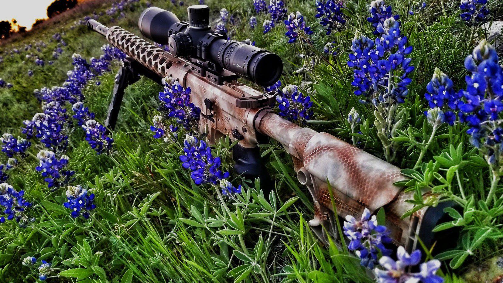 Camouflaged rifle with scope rests in bluebonnet flowers.
