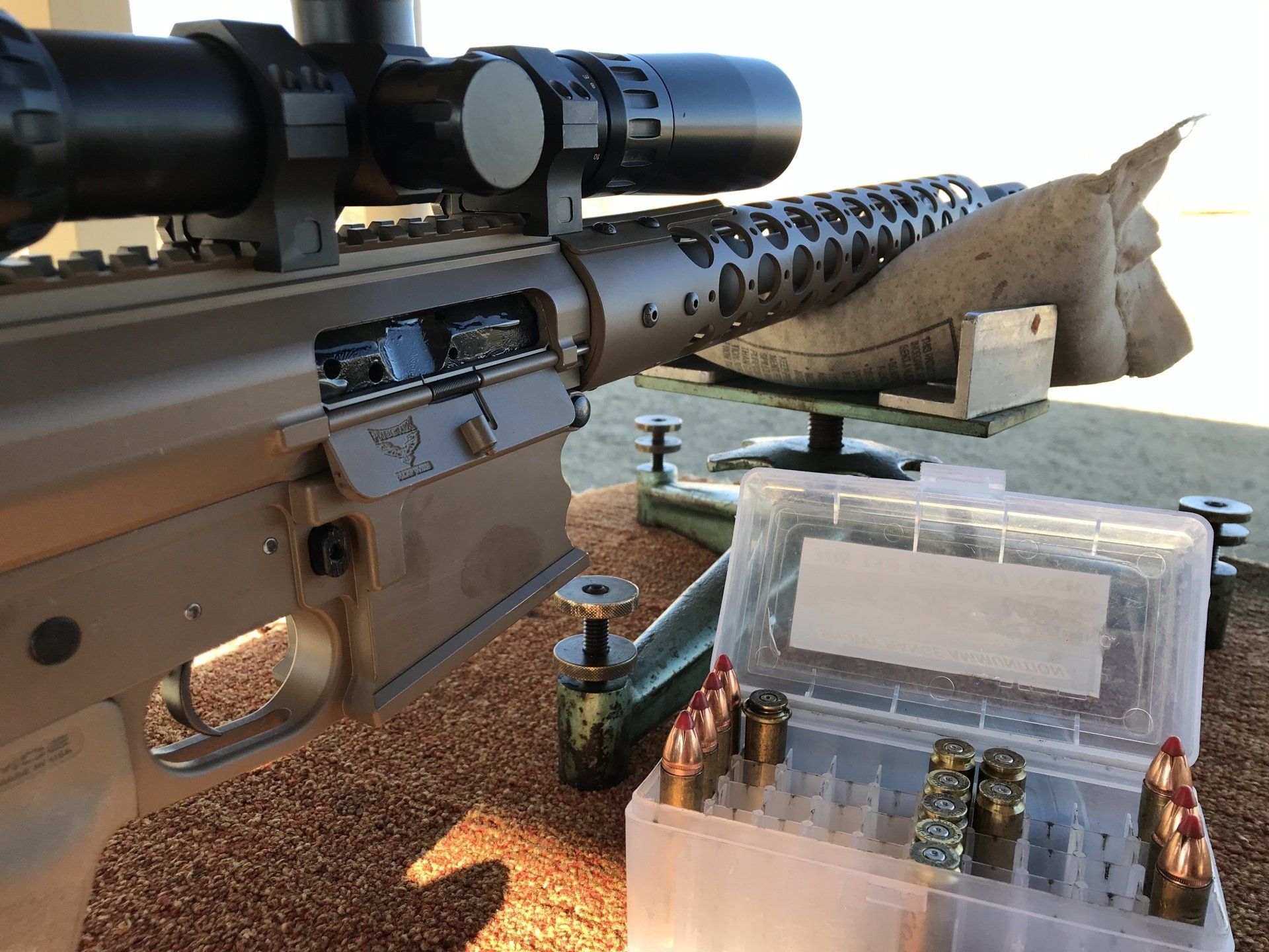 Rifle with scope on a shooting range, next to a box of ammunition.