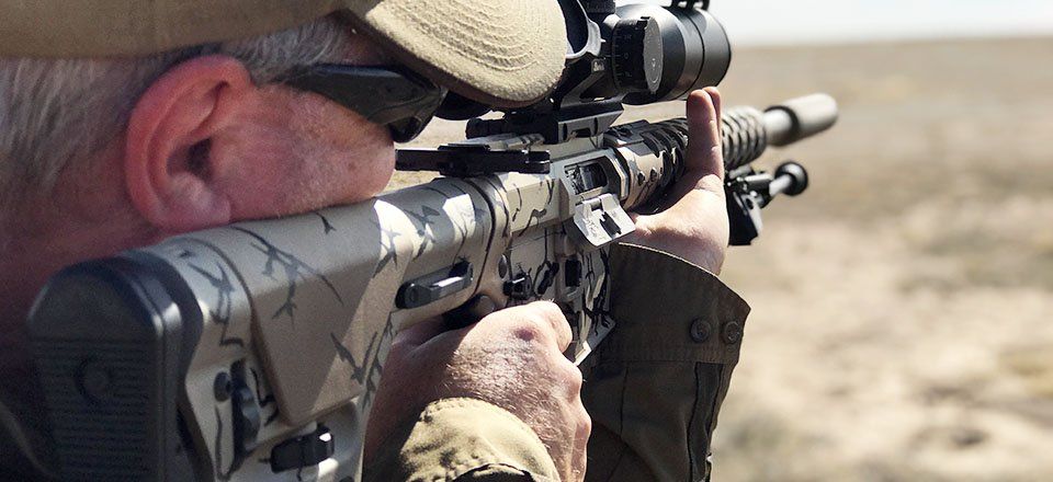 Person aiming a camouflaged rifle with a scope in an outdoor, arid environment.