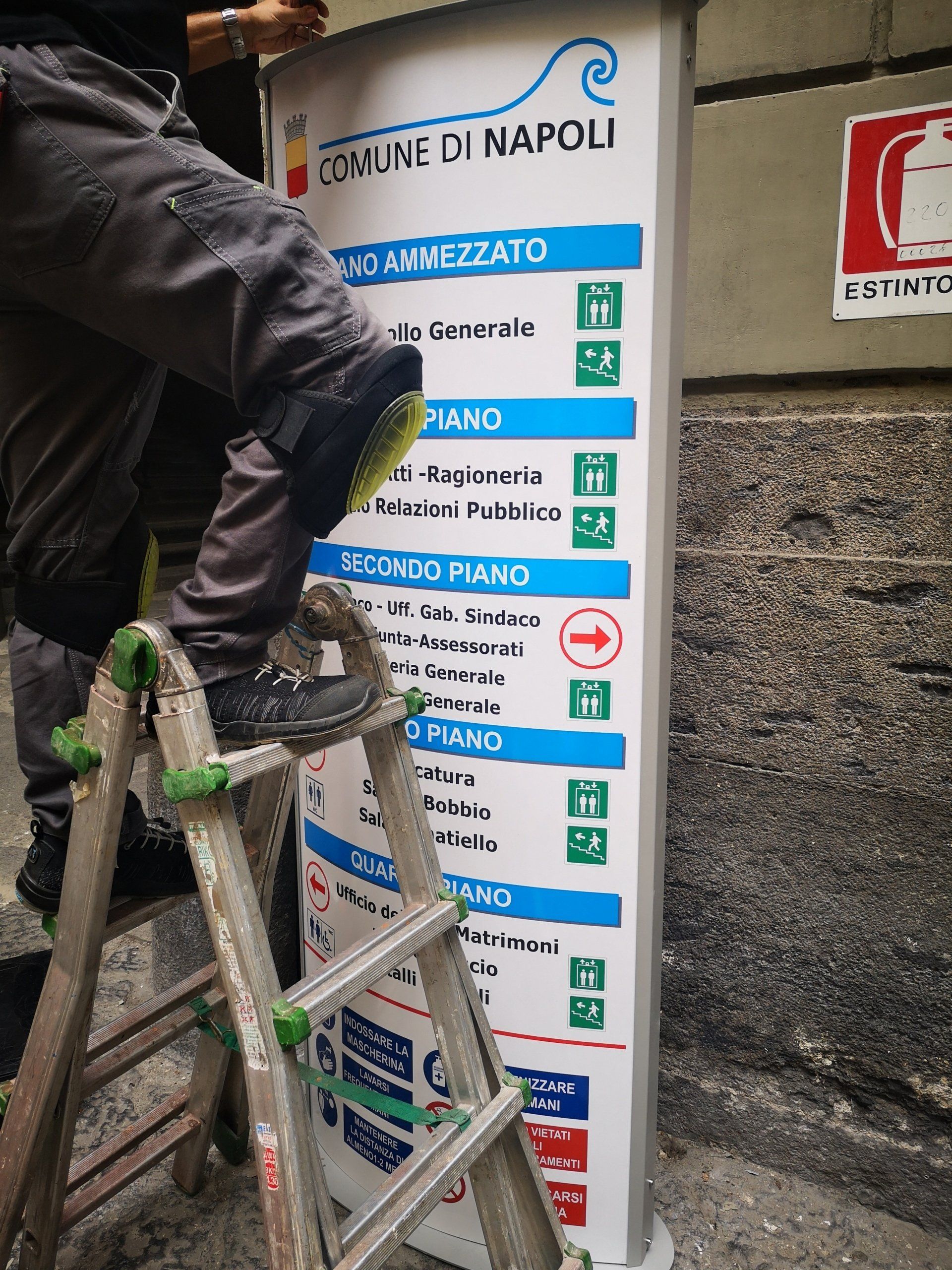 Una persona su una scala accanto a un cartello blu e bianco di Napoli, vicino a un muro di pietra.