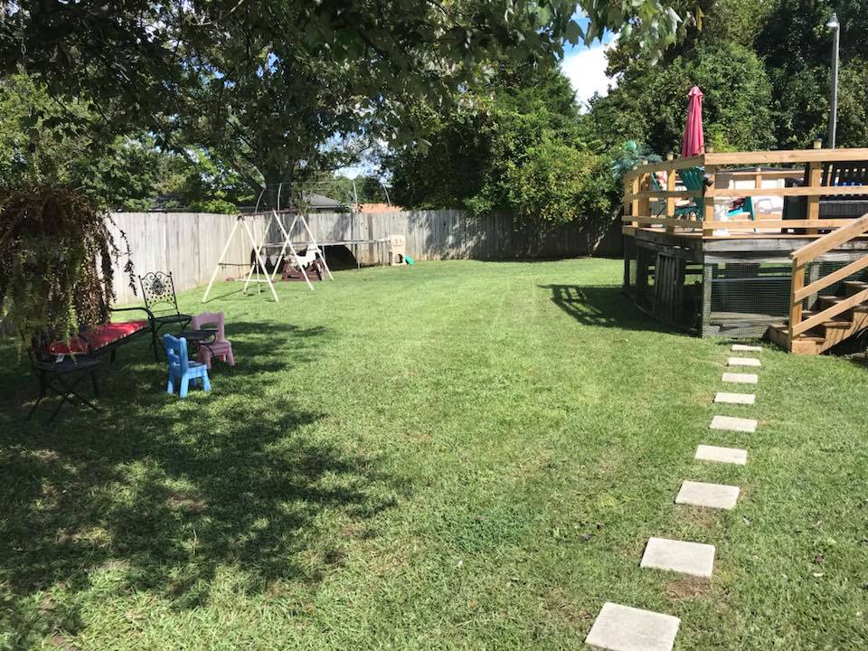 Green backyard with a wooden deck, stepping stones, swing set, and a fence.