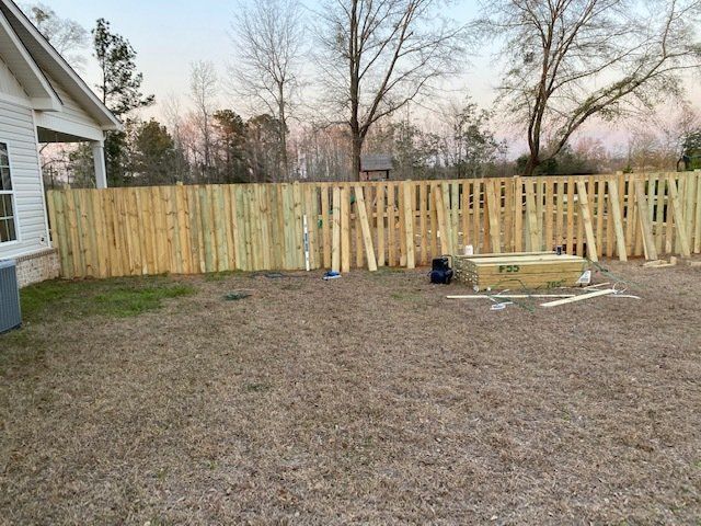 Backyard with wooden fence, brown grass, and materials; house on left, trees in background.