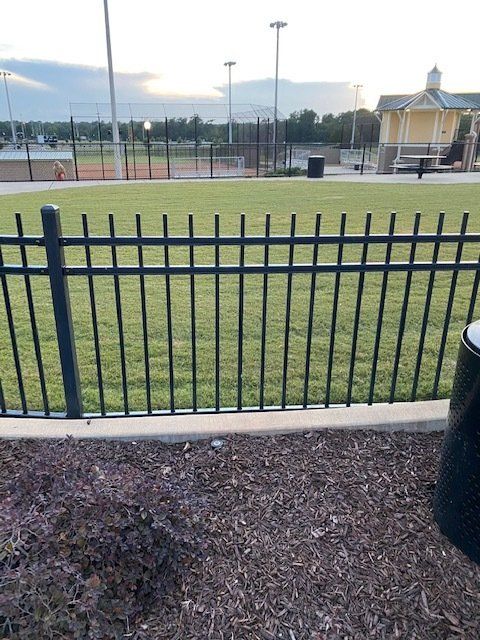 Black metal fence bordering a green grassy area with tennis courts, trees, and a pavilion in the background.