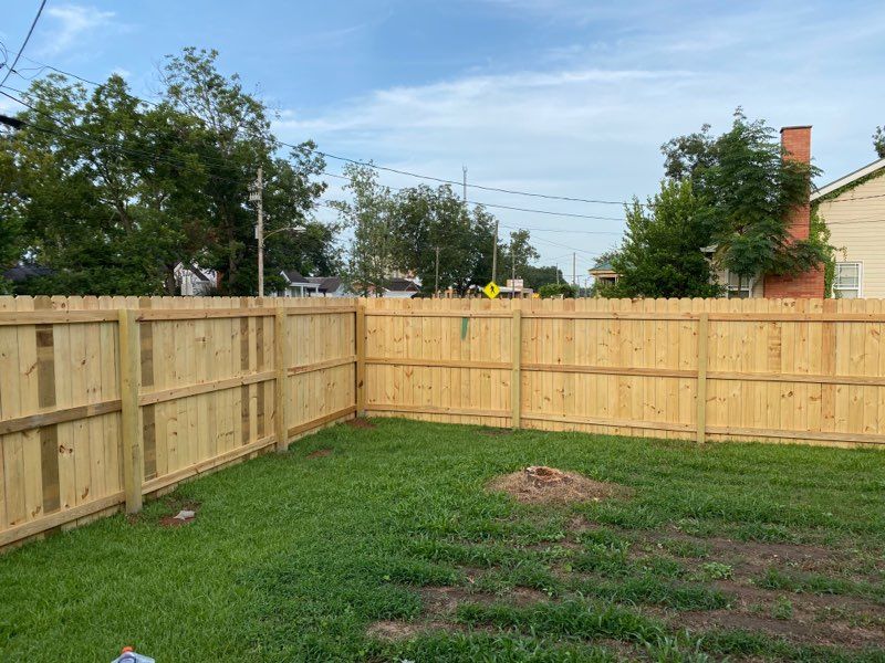 Wooden fence encloses a green lawn, with trees and a brick chimney in the background under a cloudy sky.