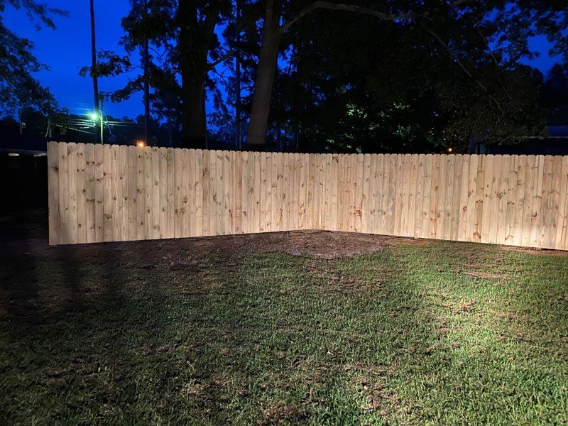 Wooden fence in a yard at night, lit by spotlights, with a dark blue sky.